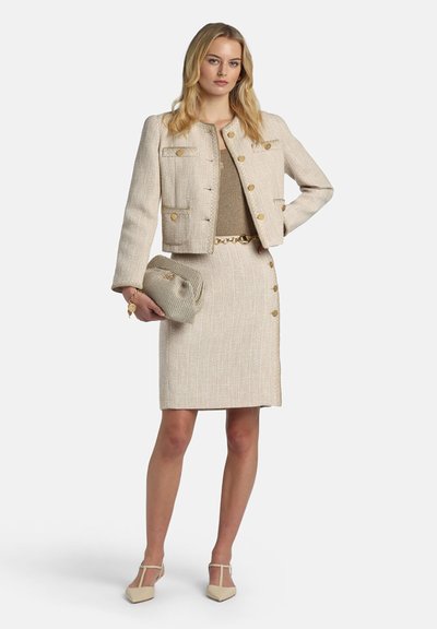 Woman wearing beige textured jacket and skirt with gold buttons, holding matching clutch and wearing beige heels, standing with hand on hip.