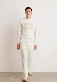Weißes Sweatshirt mit beigem "HUGO"-Logo auf der Vorderseite, kombiniert mit cremefarbenen Hosen und hellen Sneakern. Einfaches Design mit lässiger Passform.