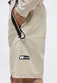 Shorts beige Berghaus à motif quadrillé, avec une poche zippée noire et une main partiellement insérée dans la poche.
