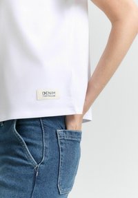 Fehér pamut ing oldalsó címkével, rajta a "DENIM TOM TAILOR" felirattal. Kék farmernadrággal párosítva, amelyen hátoldali zsebrészletek láthatók.