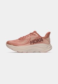 Baskets de course Hoka en rose doux, avec une tige en maille respirante, un col rembourré, une épaisse semelle blanche et un logo Hoka bien visible sur le côté.