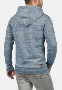 Sudadera de hombre en tejido azul jaspeado, con capucha ajustable, puños acanalados y una etiqueta de marca de cuero en la parte baja de la espalda.