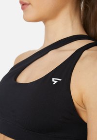 Črn športni nedrček z eleganto zasnovo, dvojni naramnici, širokim pasom in majhnim belim logotipom na levi strani. Gladka tekstura tkanine.