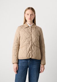 Kvinde iført en beige quiltet jakke med knaplukning og frontlommer, parret med blå jeans, stående imod en ensfarvet baggrund.