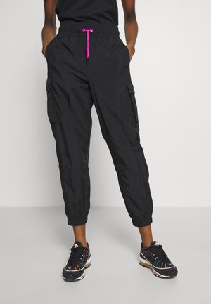 Person iført sorte cargo-joggerbukser med pink snor, kombineret med sorte sneakers med flerfarvede detaljer, stående med hænderne i lommerne.