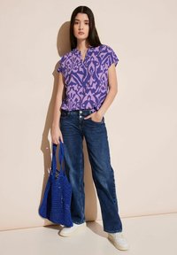 Top viola a maniche corte con un design a fantasia, abbinato a jeans blu e sneaker bianchi. Tiene in mano una borsa a tracolla blu realizzata all'uncinetto.