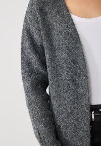 Grijze, fuzzy cardigan met een open voorkant. Kenmerkt een zachte textuur en lange mouwen, gedragen over een wit topje en zwarte broek.