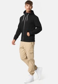 Fekete cipzáras kapucnis pulóver elülső zsebbel, állítható kapucnival és zsinórokkal, bézs cargo nadrággal és fehér magas szárú tornacipővel párosítva.