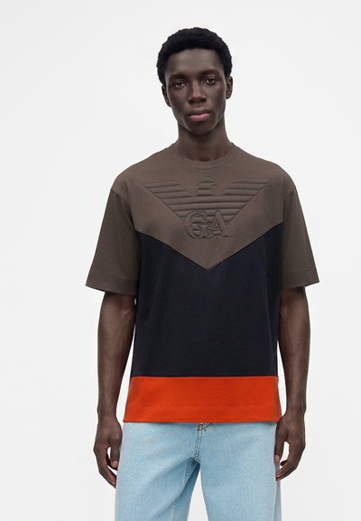 Jeune homme portant un t-shirt à manches courtes color-block avec des sections marron, noir et orange et un jean bleu clair sur un fond uni.