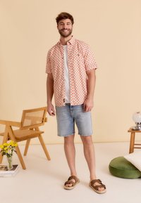 Korte mouwen, gestreept shirt in koraal en wit, gecombineerd met lichtdenim shorts. Bruine sandalen met dubbele banden. Lichte achtergrond.