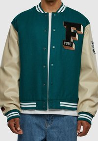 Teal vlna smíšená varsity bunda s krémovými koženými rukávy, na které je černě vyšitý symbol "F" a logo "FUBU". Hnětené manžety a pas.