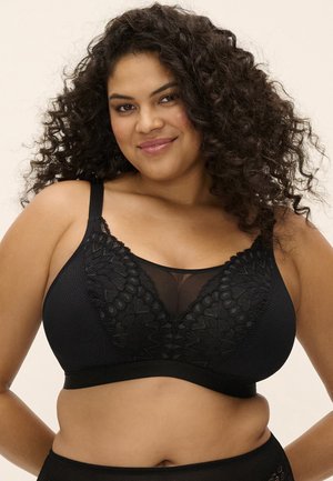 Mujer sonriente con cabello rizado que lleva un sujetador de encaje negro y ropa interior a juego sobre un fondo neutro.