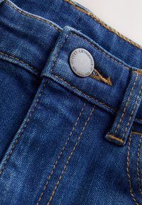 Gros plan sur un jean en denim bleu montrant un bouton métallique gravé de "Next Authentic Est. Since 1982" et des coutures brunes détaillées.
