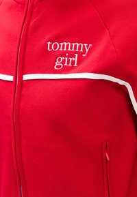 Sudadera roja con cremallera hecha de tela suave con ribetes blancos. Presenta el logo "tommy girl" en bordado blanco en el pecho. Detalle de bolsillo pequeño.