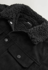 Schwarze Wildlederjacke mit einem strukturierten, flauschigen schwarzen Kragen, zwei Fronttaschen und schwarzen Druckknöpfen. Innenlabel sichtbar.
