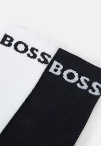 Δύο ζεύγη κάλτσες σε λευκό και μαύρο με ραφή ριμπ, featuring το λογότυπο "BOSS" υφαντό σε αντίθετα χρώματα σε καθεμία κάλτσα.