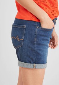 Nainen, joka tarjoaa käärittyjä farkkushortsit, joissa on oranssia ommeltu tasku, ja oranssi reikäneulepaita, käsi etutaskussa.