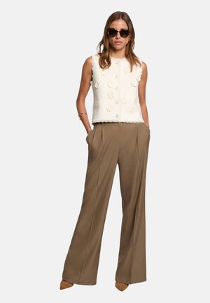 Femme portant un haut sans manches blanc orné de motifs floraux, un pantalon marron à jambes larges, des chaussures pointues marron et des lunettes de soleil foncées, debout les mains dans les poches.