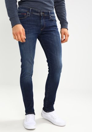 Dark blue slim-fit jeans with a smooth texture, contrast stitching, five-pocket design, and a mid-rise waist. Paired with white sneakers.