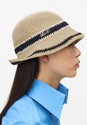 Bočni profil osobe koja nosi bež kukičanu kačket kapu s crnim i bijelim prugama te plavu košulju s ovratnikom.