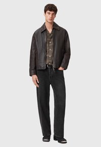 Giacca in pelle marrone con colletto, indossata sopra una camicia a scacchi con bottoni. Modello abbinato a jeans larghi neri e stivali neri.