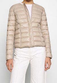 Giacca a vento beige, design trapuntato con linee orizzontali strette, senza zip sul davanti, tessuto leggero, abbinata a pantaloni bianchi a vita alta.