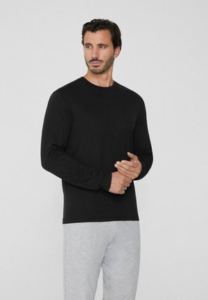Homme avec des cheveux courts et foncés et une barbe, portant une chemise noire à manches longues et un pantalon gris clair, se tenant contre un fond clair uni.