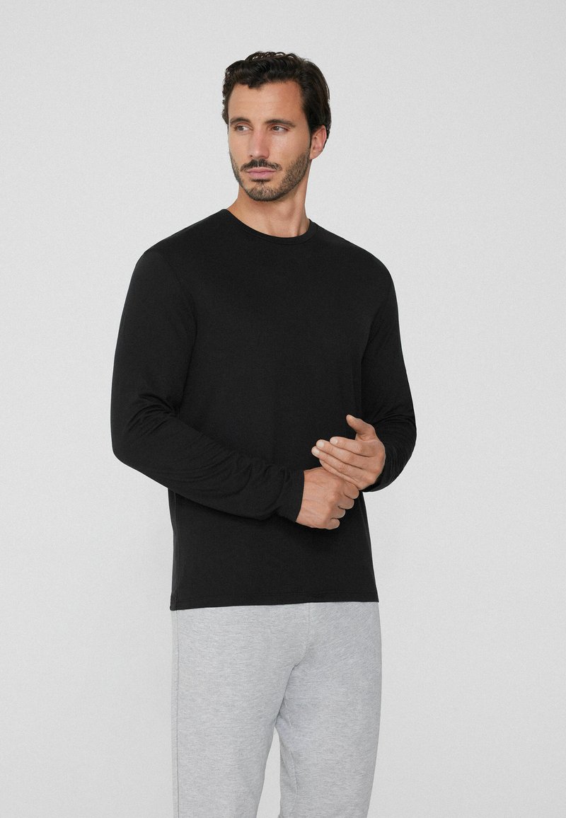 Mann mit kurzen dunklen Haaren und Bart, der ein schwarzes Langarmshirt und hellgraue Hosen trägt und vor einem einfarbigen hellen Hintergrund steht.