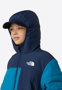 Φουσκωτό μπουφάν με συνδυασμό χρωμάτων ναυτικού μπλε και τιρκουάζ, φερμουάρ μπροστά, κουκούλα και λεπτομέρεια λογότυπου. Το ύφασμα φαίνεται λείο και μονωμένο.