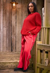 Femme portant un pull en tricot rouge et un pantalon large rouge à rayures blanches, debout sur des marches en bois à côté d'une rampe sous une lumière chaude.