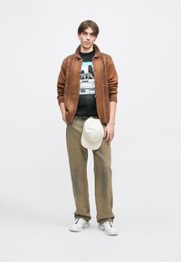 Καφέ ριγέ ανοιχτό πουλόβερ πάνω από μαύρο γραφικό T-shirt, ανοιχτό πράσινο παντελόνι, λευκά αθλητικά παπούτσια και κρέμα καπέλο κρατημένο στο χέρι. Απλή φόντο.