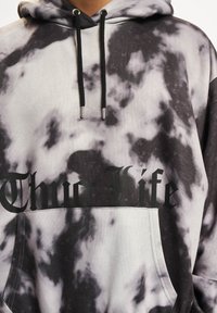 Črno-bele tie-dye kapuce s sprednjo žepom, ki nosi besedilo "Thug Life" v krepki črni pisavi. Tkanina ima mehak občutek in se prilagaja z vrvicami.