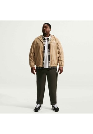 Mann steht und trägt eine beige Kapuzenjacke über einem schwarz-weiß karierten Hemd, dunkle Hosen und schwarz-weiße Sneakers vor schlichtem Hintergrund.