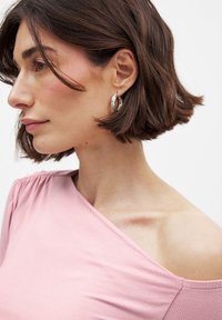Roze off-shoulder top van zacht materiaal, met een geplooid detail aan één kant, aangevuld met zilveren ringoorbellen.