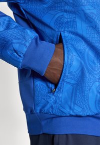 Modrá mikina s textúrovaným vzorom s písmenami, so zipsovou vreckom na boku a rebrovanými manžetami pre priliehavé nosenie. Hladký povrch látky.