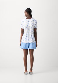 Camicetta bianca a maniche corte con motivo floreale blu, abbinata a pantaloni corti azzurri. Presenta una schiena arricciata e una vestibilità ampia. Tacchi trasparenti.