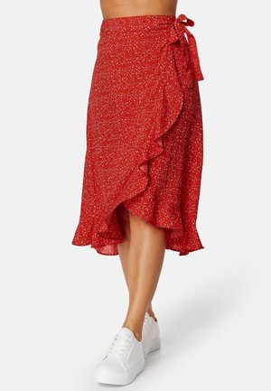Jupe portefeuille rouge à pois blancs, bords volantés, nouée à la taille, associée à des baskets blanches compensées sur jambes nues.