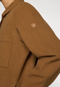 Bruine blouse met lange mouwen van gestructureerde stof, voorzien van een borstzak en een klein rond logo-patch op de linkermouw.