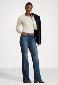 Maglia beige a maniche lunghe, jeans blu a zampa d'elefante con leggera scoloritura, cintura in pelle marrone, modello che tiene una giacca blu navy sulla spalla, sfondo neutro.