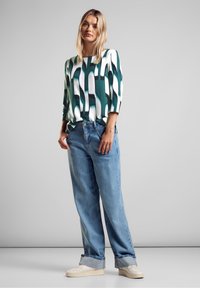 Blusa verde e bianca a fantasia con maniche a tre quarti, abbinata a jeans azzurri a vestibilità ampia e sneakers bianche.