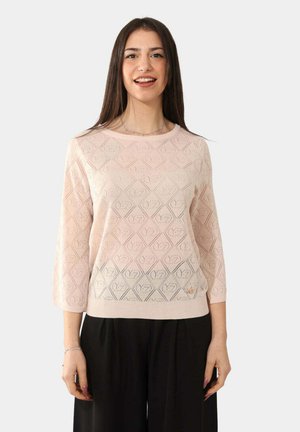 Donna che indossa una maglia a maniche lunghe rosa chiaro con fantasia e pantaloni neri larghi, sorridente, in piedi davanti a uno sfondo bianco uni.