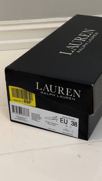 Juoda stačiakampė batų dėžutė su baltais užrašais „LAUREN RALPH LAUREN.“ Etiketėje nurodytas produkto pavadinimas (Lolah II PM SLG), ES dydis 38 ir brūkšniniai kodai.
