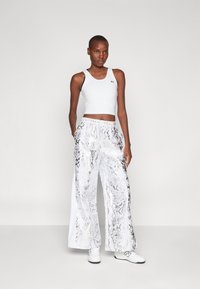 Biely športový crop top s čiernymi akcentmi, spárovaný s lesklými, širokými striebornými nohavicami s hadím vzorom a bielymi teniskami.