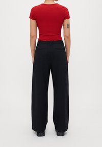 Osoba w dopasowanej, czerwonej koszulce z krótkim rękawem i czarnych, szerokich spodniach na wysokim stanie, stojąca tyłem.