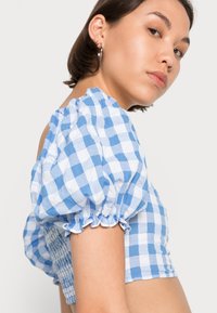 Top cropped em azul e branco com padrão de quadrados e mangas bufantes. Apresenta babados franzidos nas mangas e detalhes franzidos nas costas. Feito de tecido de algodão.