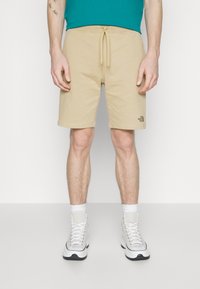 Pantaloni corti beige con fascia elastica in vita e laccio, dotati di un piccolo logo nero. Modello abbinato a sneaker bianche e calze corte.