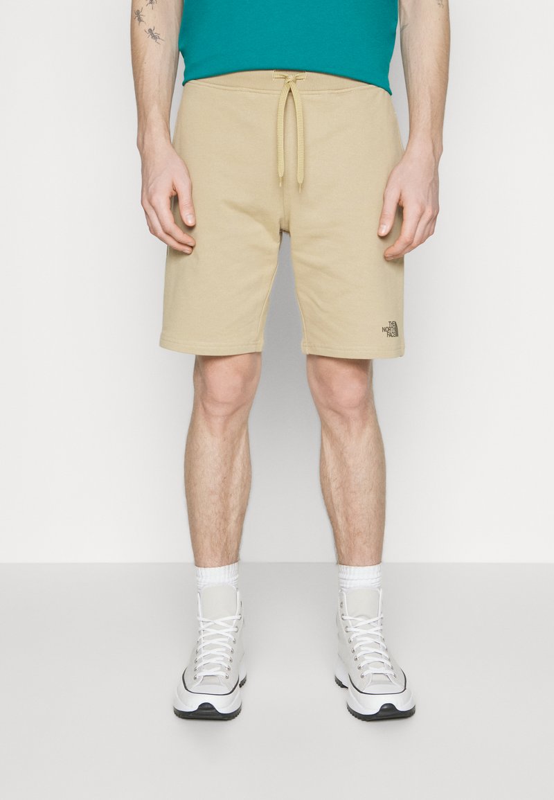 Pantaloni corti beige con fascia elastica in vita e laccio, dotati di un piccolo logo nero. Modello abbinato a sneaker bianche e calze corte.