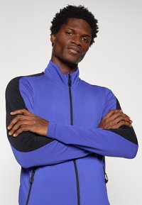 Zila sporta jaka ar augstu apkakli, melnām garajām piedurknēm, ar rāvējslēdzēju un sānu kabatām, izgatavota no gludas, elastīgas auduma.