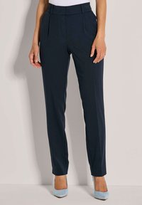 Vrouw in een nauwsluitende marineblauwe broek en lichtblauwe puntige hakken staat tegen een effen muur.