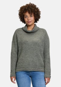 Grüner Strickpullover mit hohem Kragen, gerippter Textur, überschnittenen Schultern und langen Ärmeln, kombiniert mit blauen Denim-Jeans.